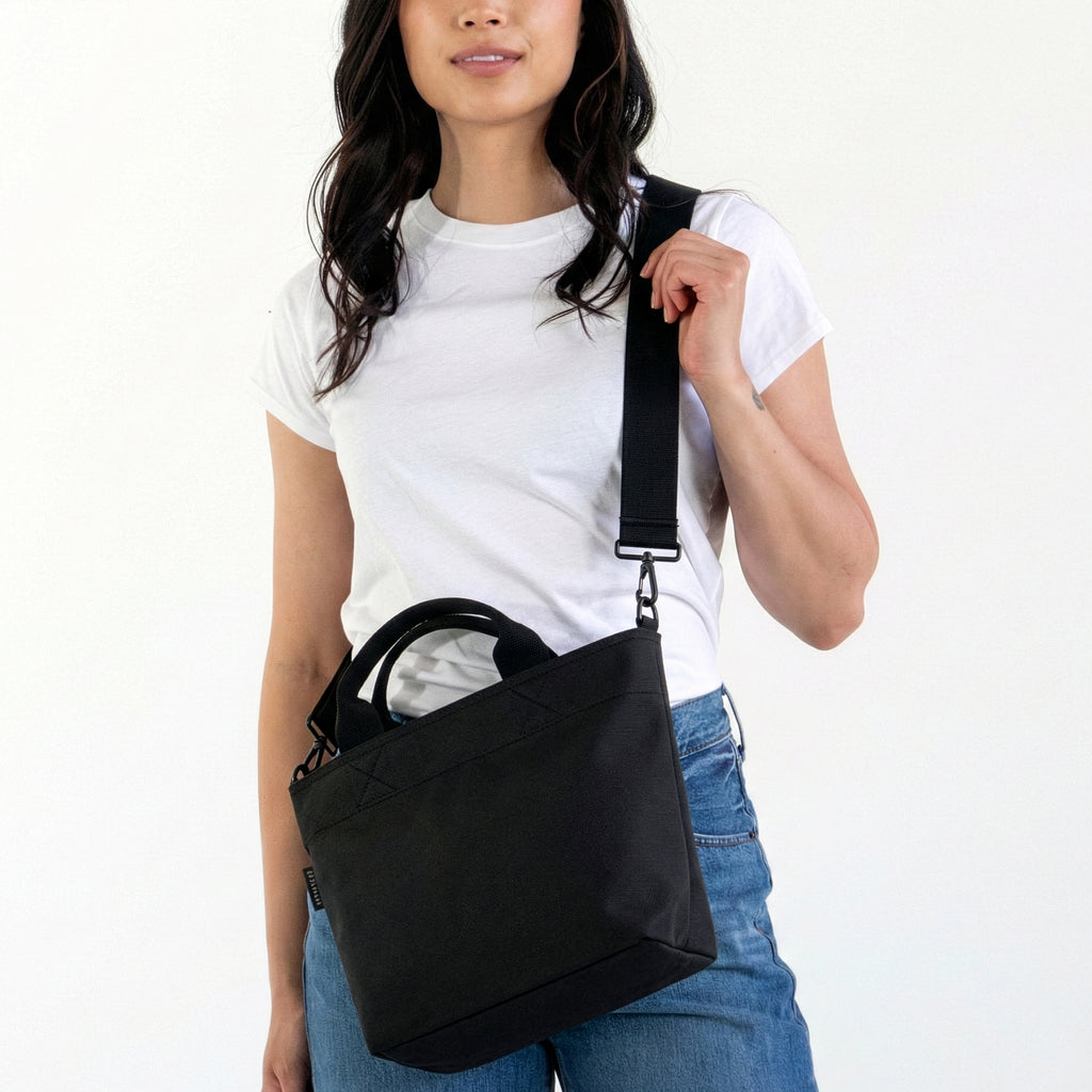Woman wearing small Canvelle Zip Crossbody Tote in black canvas with black hardware styled with a white t-shirt to demonstrate compact purse size.