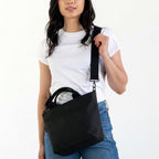 Woman wearing small Canvelle Zip Crossbody Tote in black canvas with black hardware styled with a white t-shirt to demonstrate compact purse size.