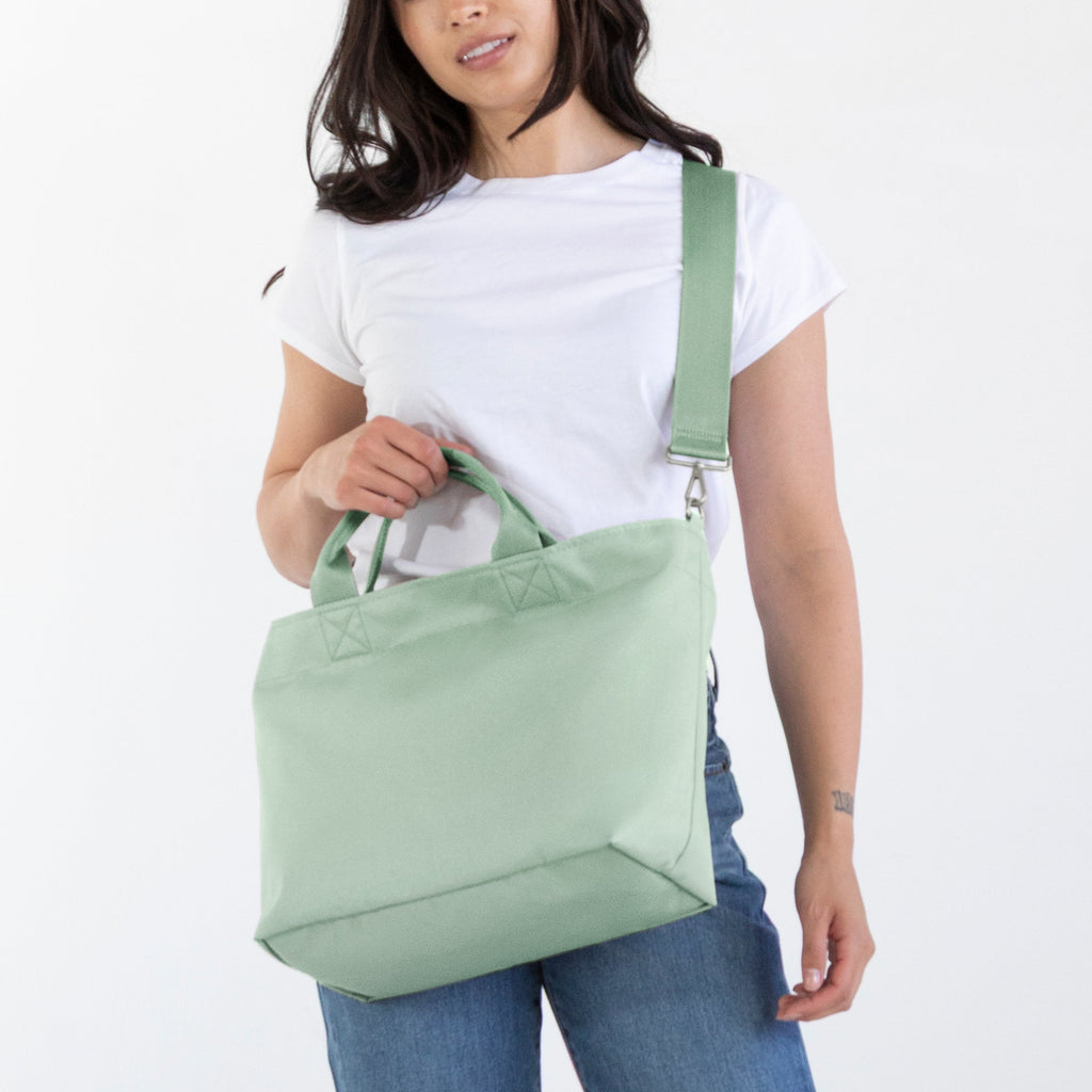 A woman modeling a sage green canvas shoulder tote bag, wearing a white t-shirt, blue jeans, and brown boots against a plain white studio background.