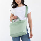 A woman modeling a sage green canvas shoulder tote bag, wearing a white t-shirt, blue jeans, and brown boots against a plain white studio background.