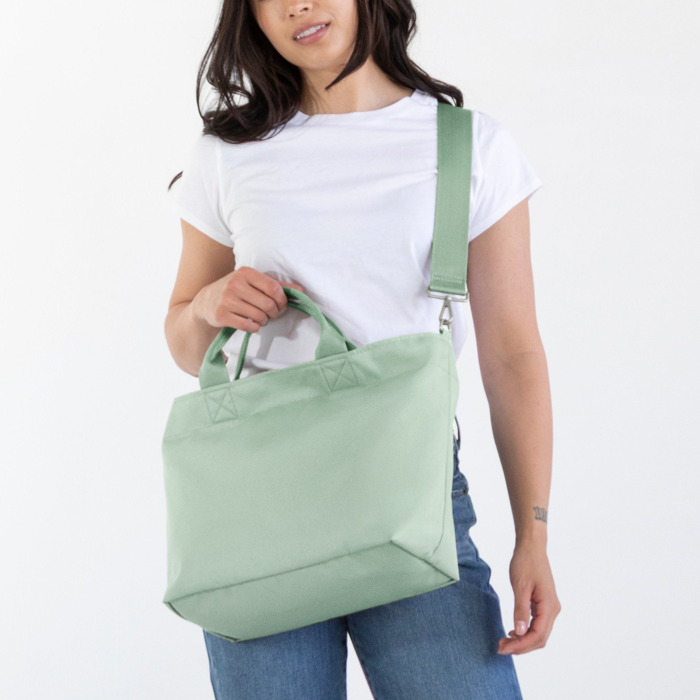 A woman modeling a sage green canvas shoulder tote bag, wearing a white t-shirt, blue jeans, and brown boots against a plain white studio background.