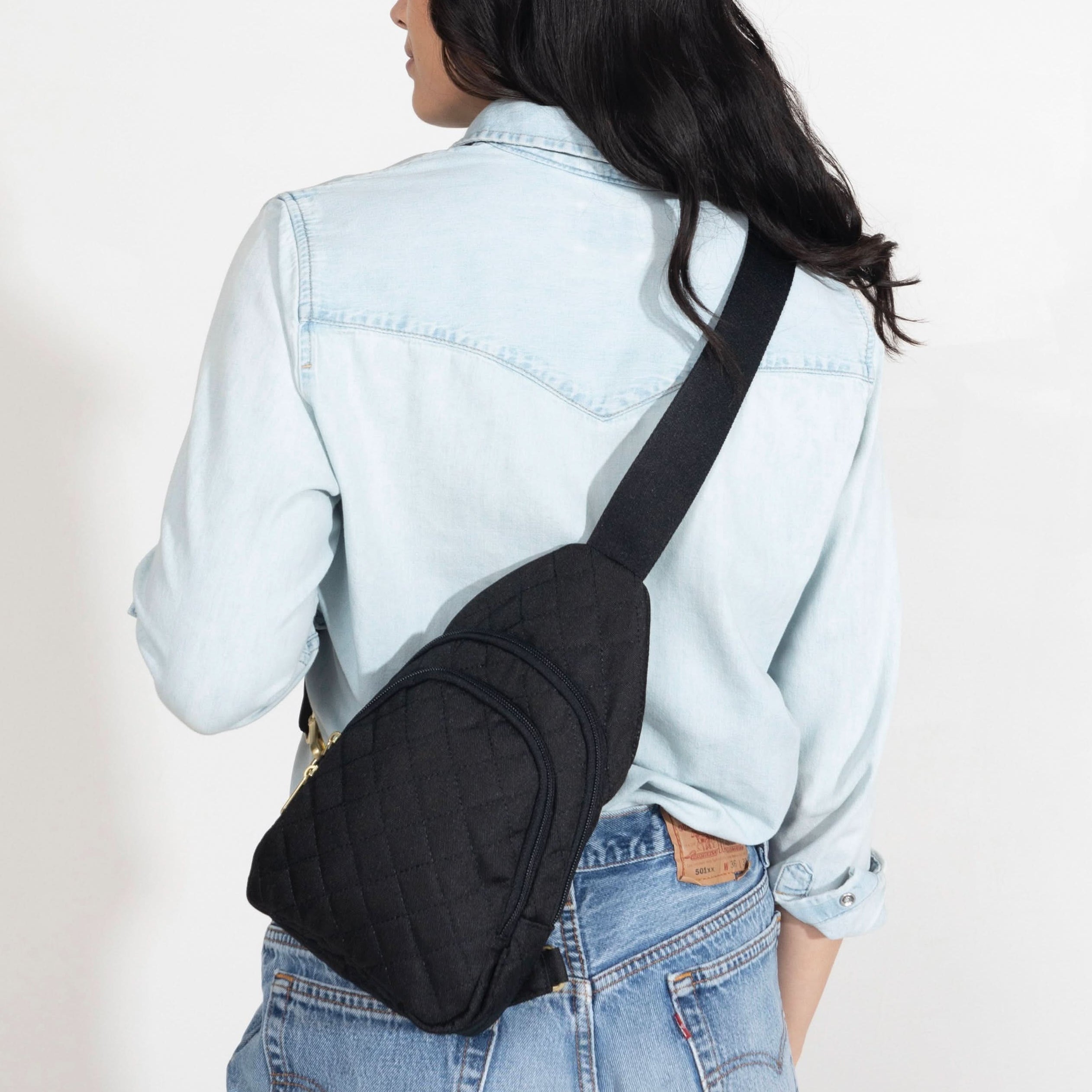 Woman wearing small Canvelle City Sling Bag in quilted black across the back over a denim shirt to demonstrate hands-free fit and textured style.