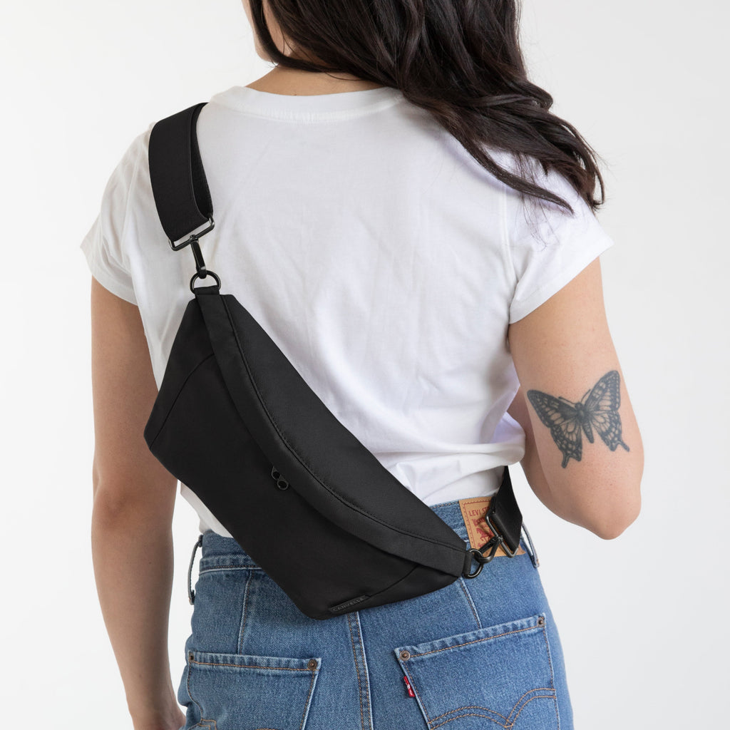 A woman viewed from behind wearing a black crossbody sling bag across her back, paired with a white t-shirt and blue jeans, against a plain white background.