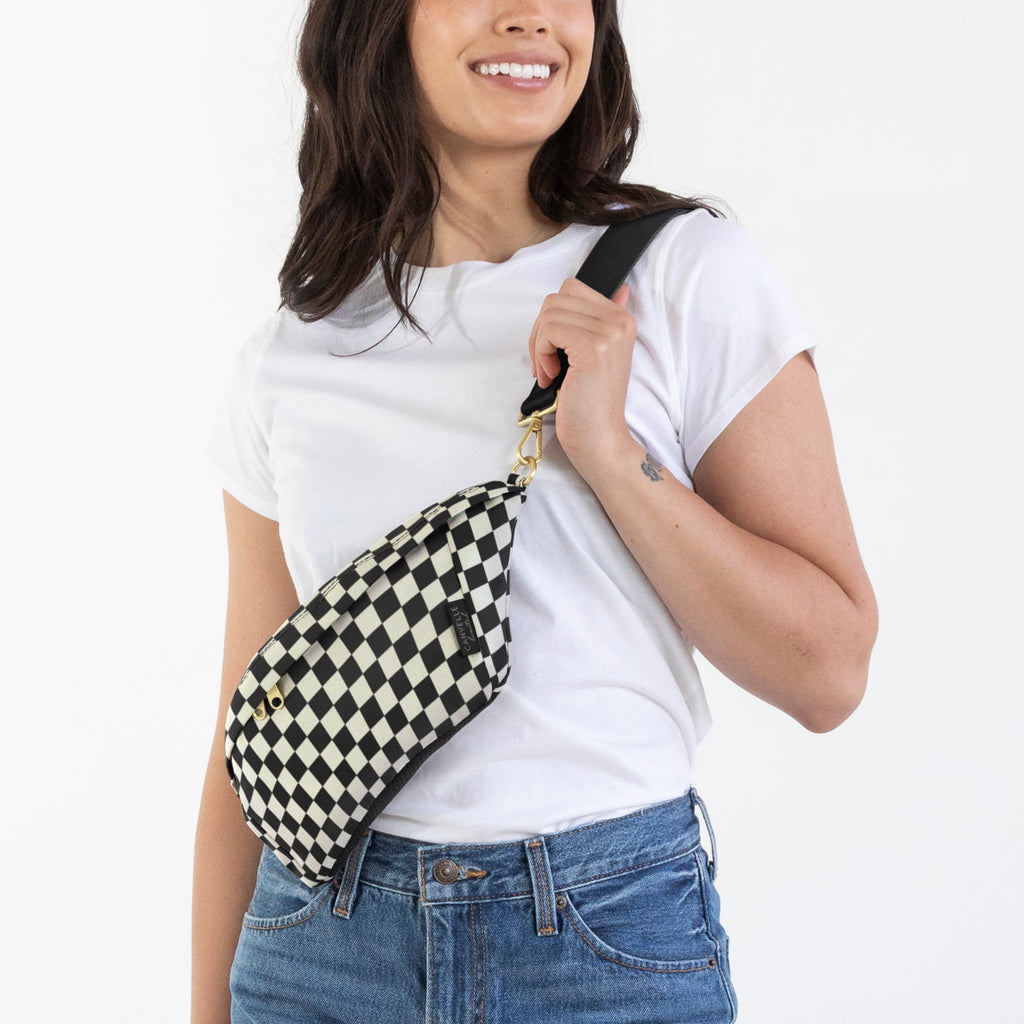 A woman modeling a black and white checkered sling bag worn crossbody over a white t-shirt and blue jeans, standing against a plain white studio background.