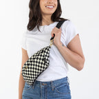 A woman modeling a black and white checkered sling bag worn crossbody over a white t-shirt and blue jeans, standing against a plain white studio background.