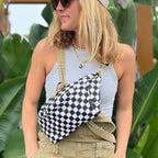 A woman wearing a beige baseball cap, sunglasses, a gray tank top, and khaki overalls carries a black and white checkered crossbody bag on her shoulder, set against a background of large green tropical leaves.