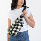 A woman modeling a black and white checkered crossbody oversized fanny pack bag, styled with a white t-shirt, blue jeans, and brown boots against a plain white studio background.