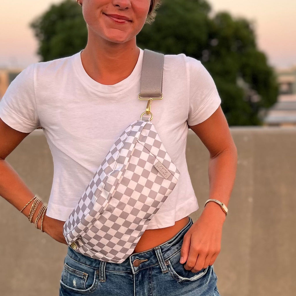 A person outdoors wearing a white and gray checkerboard sling bag across their torso, paired with a white t-shirt and blue jeans.