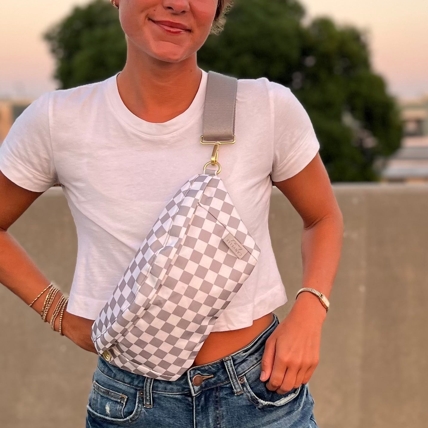 A person outdoors wearing a white and gray checkerboard sling bag across their torso, paired with a white t-shirt and blue jeans.