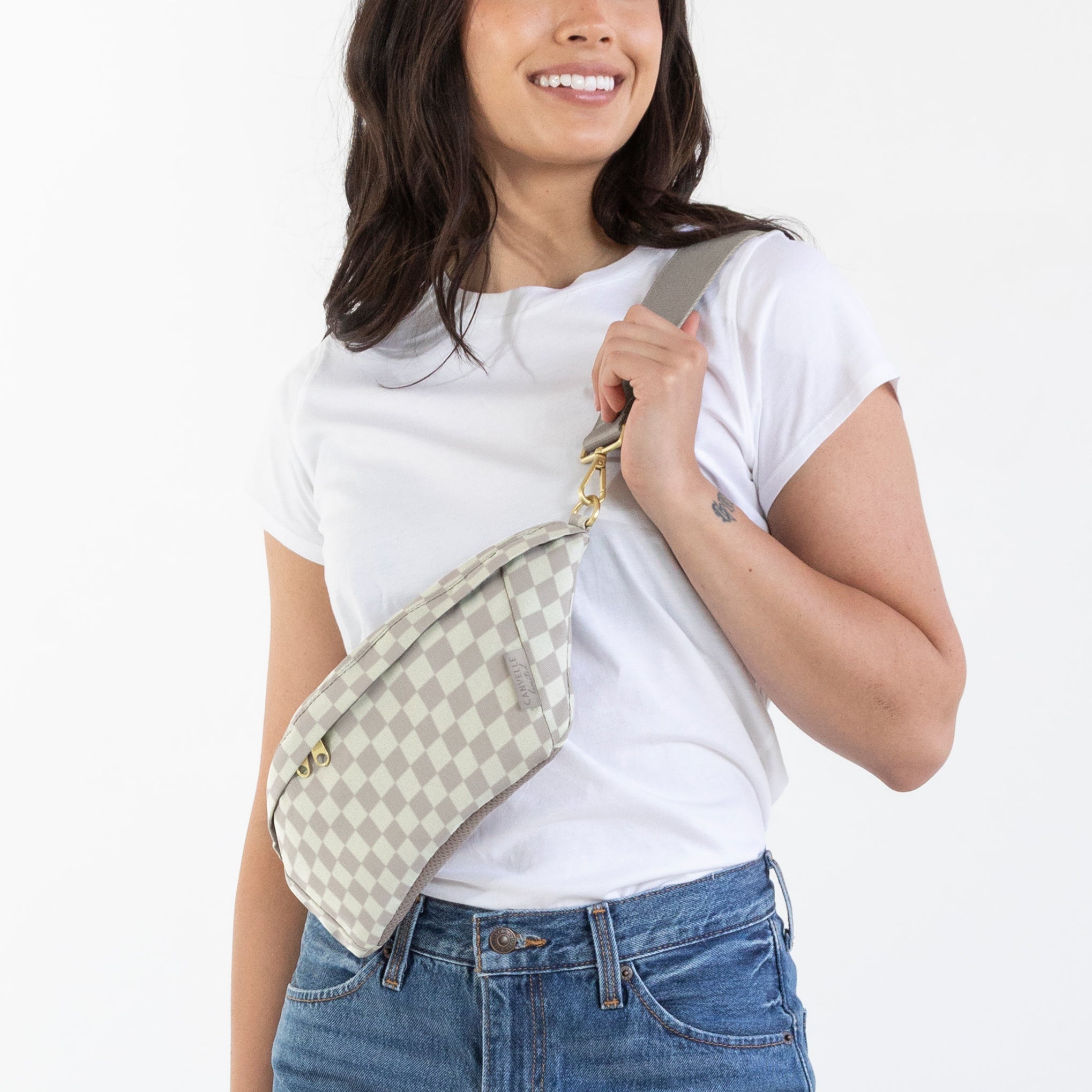 A woman modeling a white t-shirt, blue jeans, and brown boots, carrying a beige and white checkered shoulder bag against a plain white background.