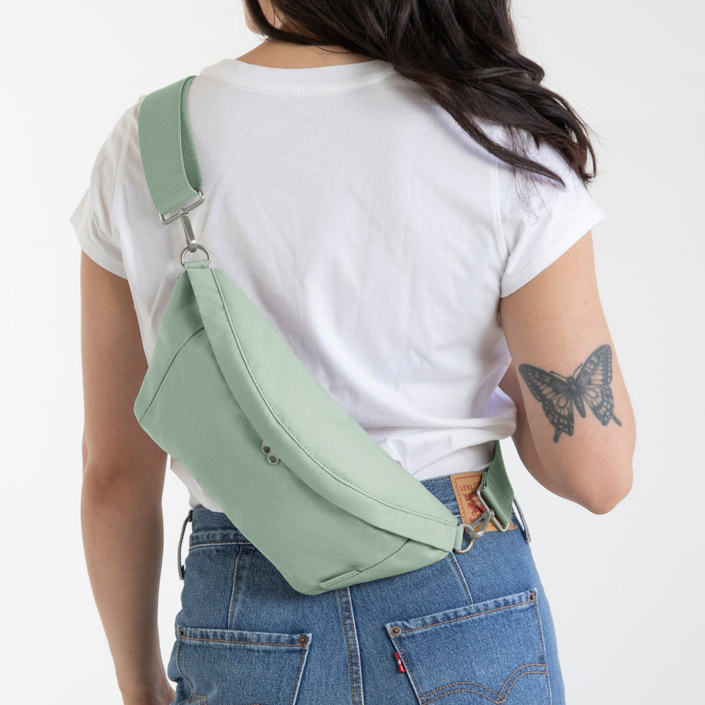 A woman viewed from behind wearing a mint green crossbody bag across her white t-shirt and blue jeans, against a plain white background.