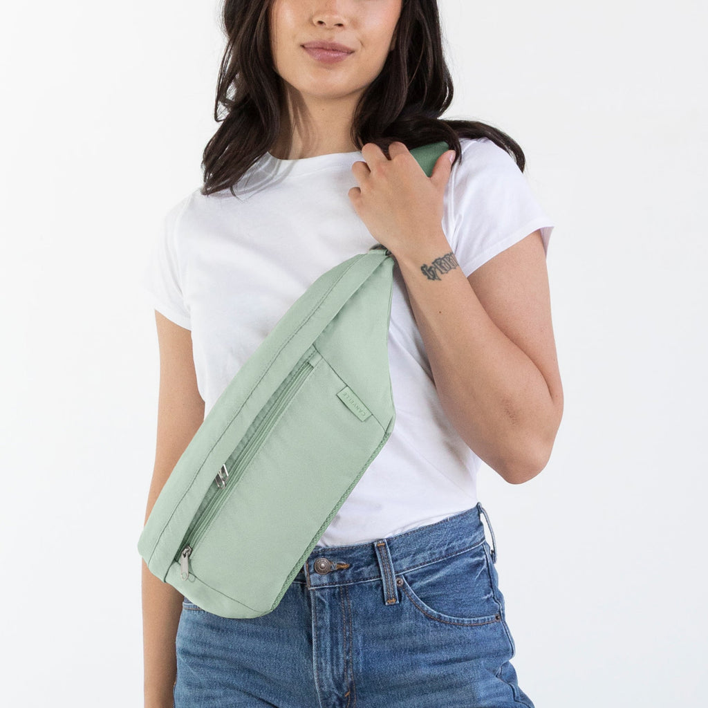 A woman modeling a light sage green envelope-style shoulder bag, paired with a white t-shirt, blue jeans, and brown leather boots, standing against a plain white studio background.