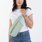 A woman modeling a light sage green envelope-style shoulder bag, paired with a white t-shirt, blue jeans, and brown leather boots, standing against a plain white studio background.