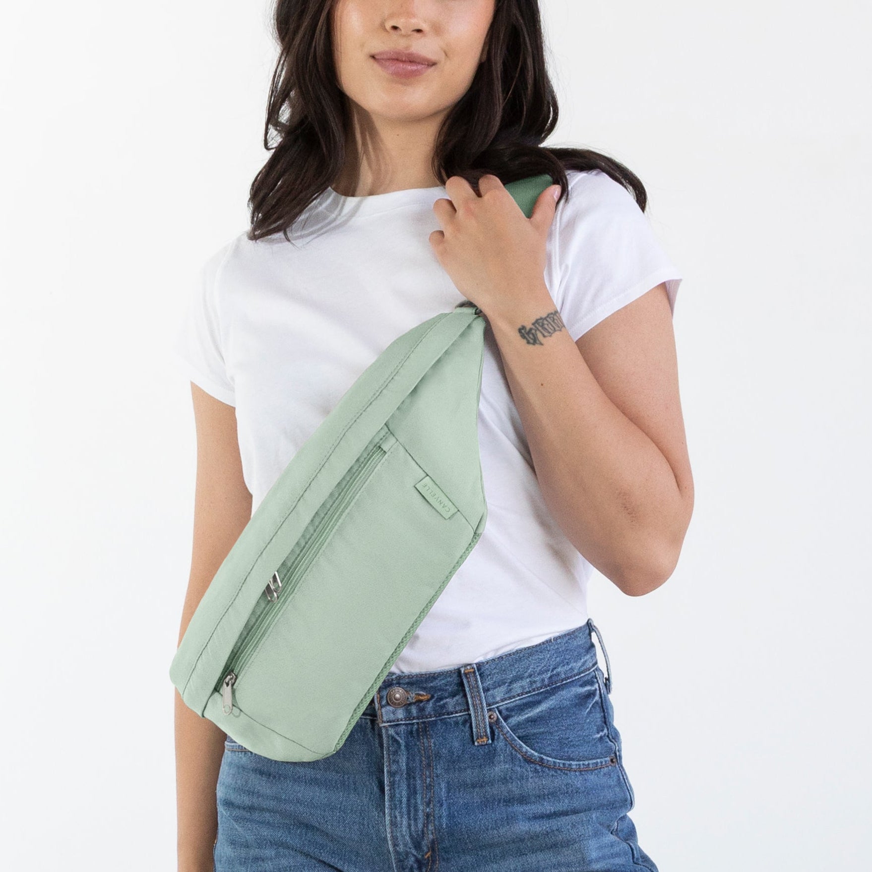 A woman modeling a light sage green envelope-style shoulder bag, paired with a white t-shirt, blue jeans, and brown leather boots, standing against a plain white studio background.