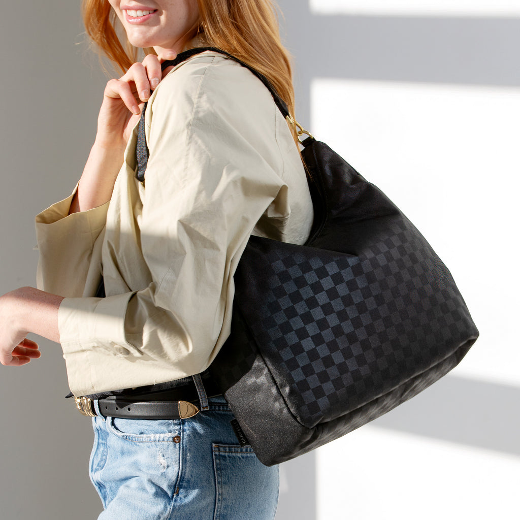 Woman wearing the large black checkered Canvelle Hobo Handbag on the shoulder to show everyday size and casual style.