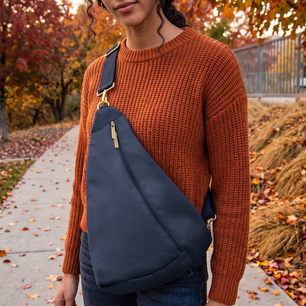 A navy blue sling bag with gold-tone hardware and a matching navy strap, worn crossbody over the chest of a person wearing an orange knit sweater in an outdoor autumn setting.