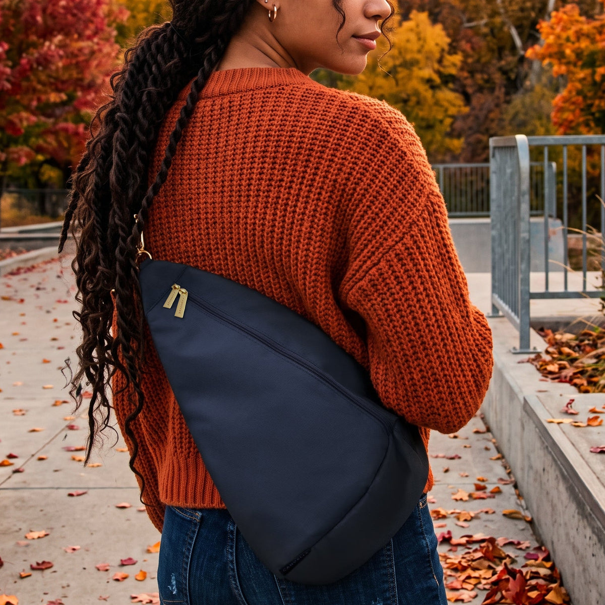 A woman with long black braids stands outdoors in an autumn park wearing a dark navy sling shoulder bag across her body. She is wearing an orange knit sweater and blue jeans, with colorful fall trees and hills in the background.