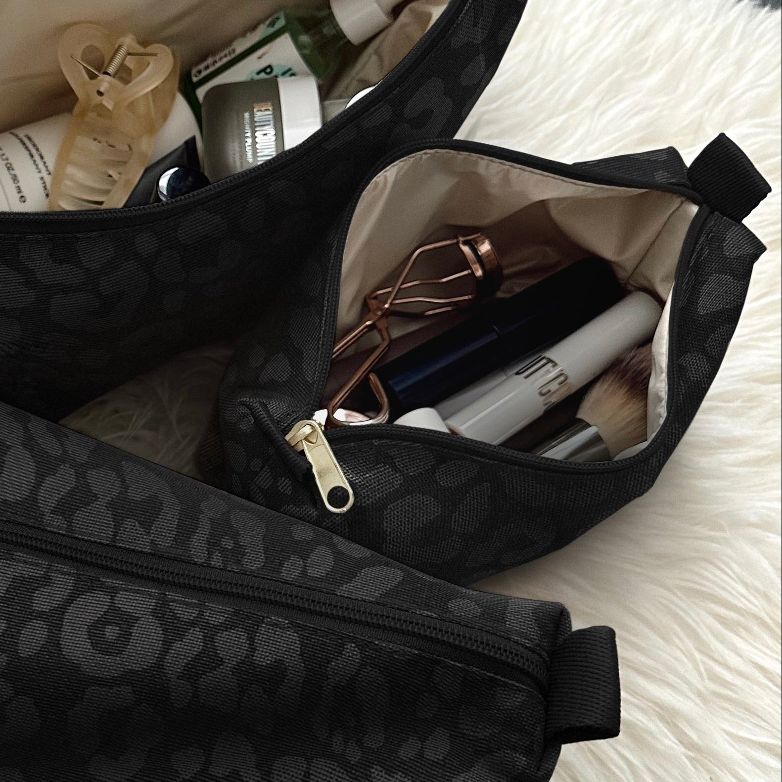 Three black leopard-print cosmetic pouches with gold zippers, shown open and filled with toiletries, arranged on a white faux-fur rug over a gray tiled floor.