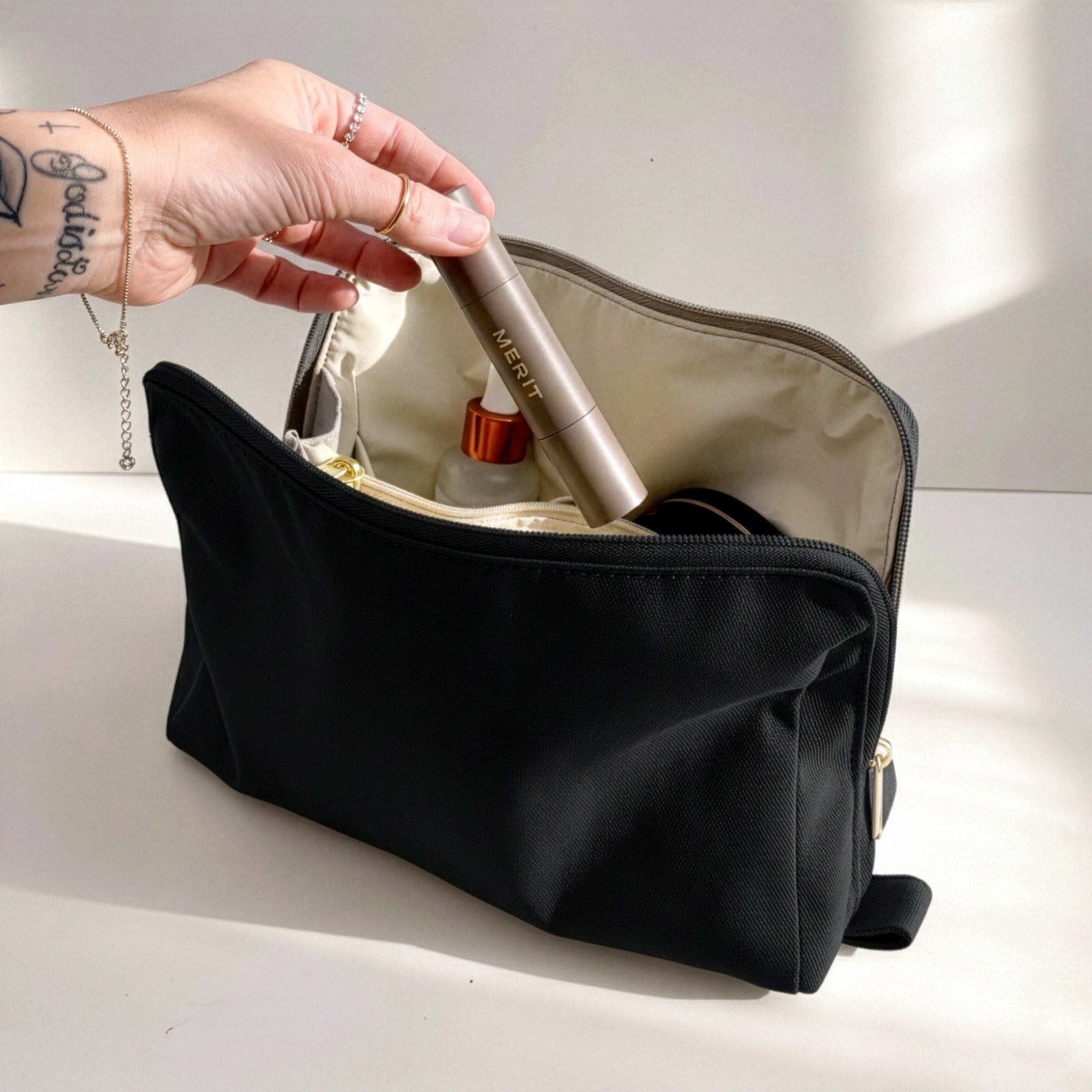 Woman's hand placing Merit makeup stick into medium black Canvelle Toiletry Bag to demonstrate compact size for daily cosmetics.